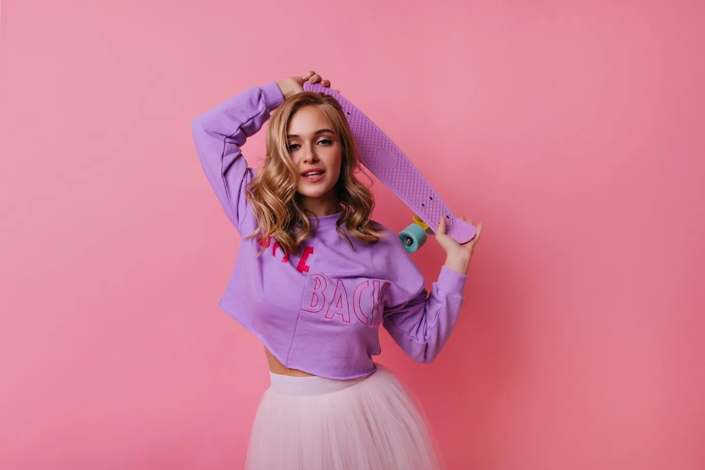 Hübsches blondhaariges Mädchen hält ein lilafarbenes Longboard. Kaukasisches weibliches Model steht im Hellen.