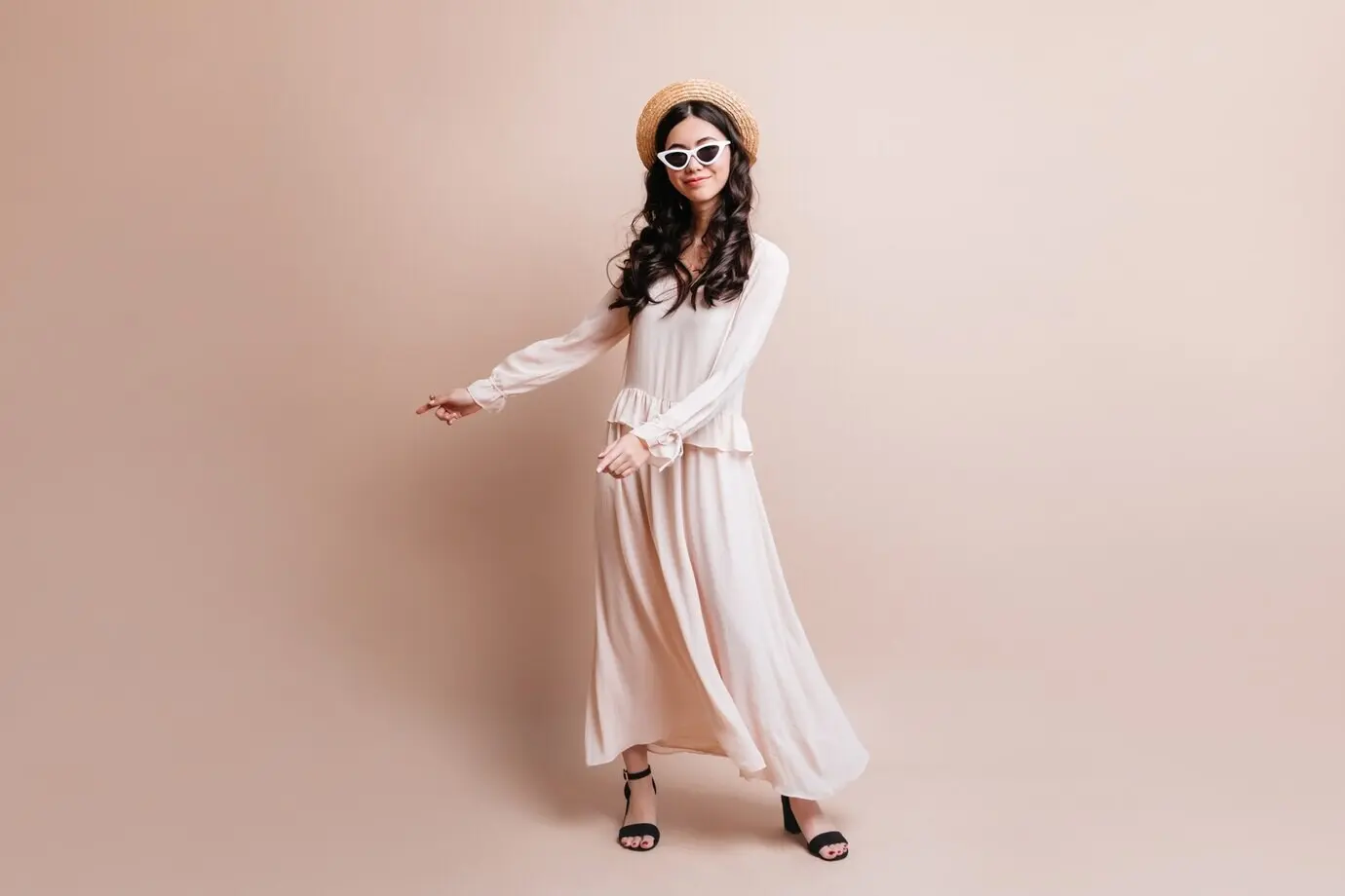 Ganzkörperansicht einer eleganten asiatischen Frau mit Hut. Studioaufnahme einer modischen japanischen Frau, die vor einem beigen Hintergrund steht.